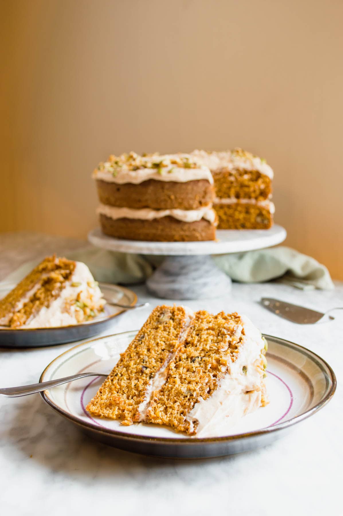 Slices of Carrot Cake with Chai Spiced Frosting on plates and cake on cake stand in background.