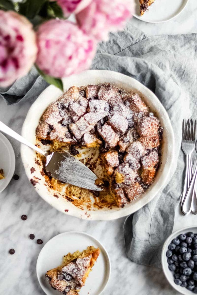 Chocolate Chip Brioche Bread Pudding in a white Emile Henry pie dish with pink peonies and a silver pie server.