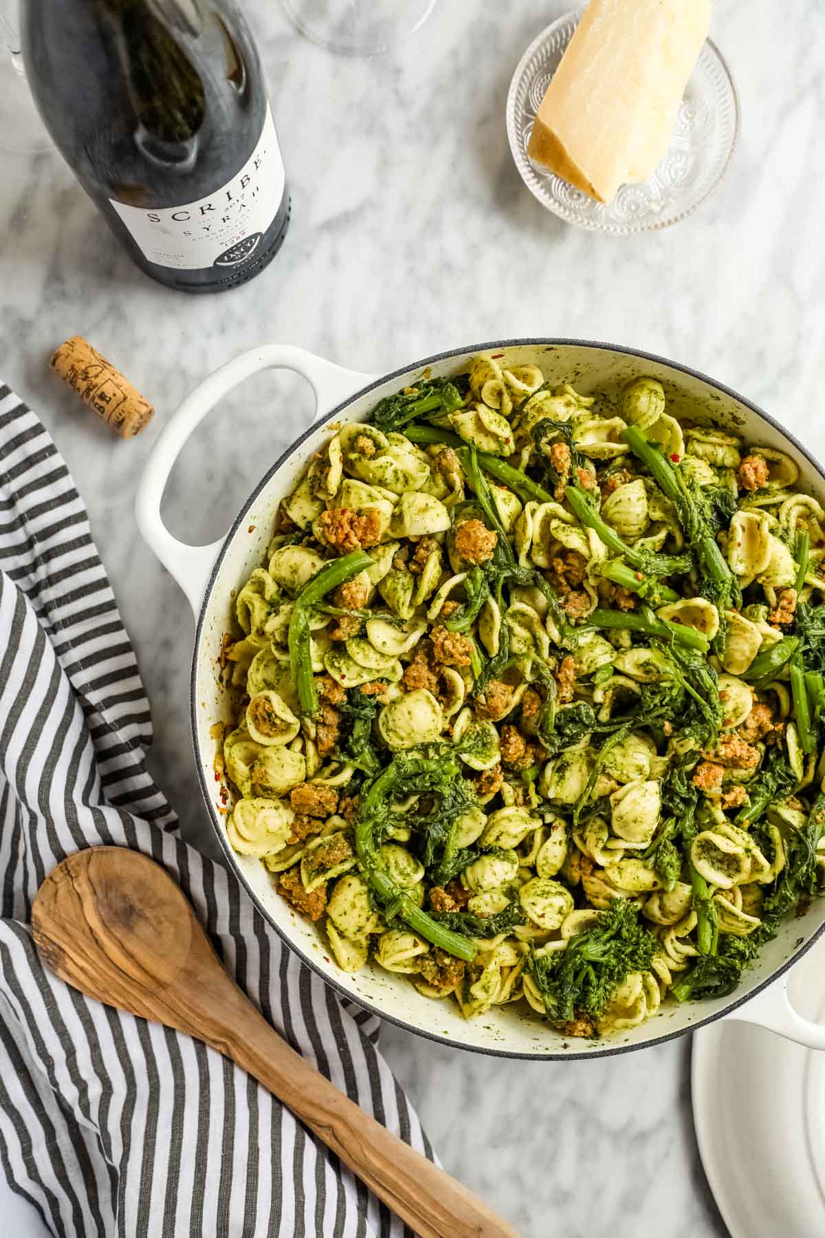 Sausage Pesto Pasta with Rapini in white braiser with wooden spoon, wine, and Parmiggianno-reggiano flatlay