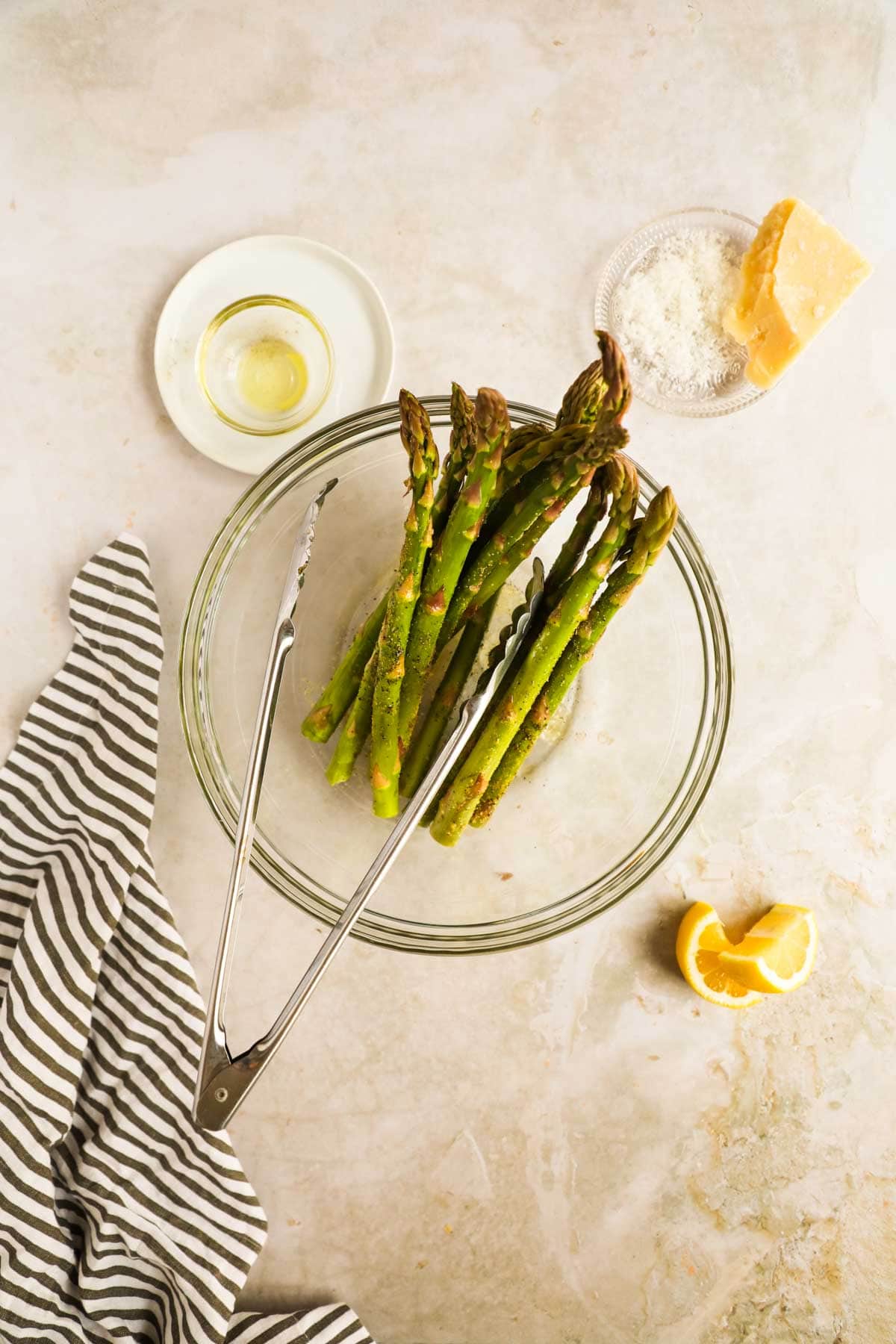 Asparagus with olive oil and seasoning in glass bowl with tongs flatlay.