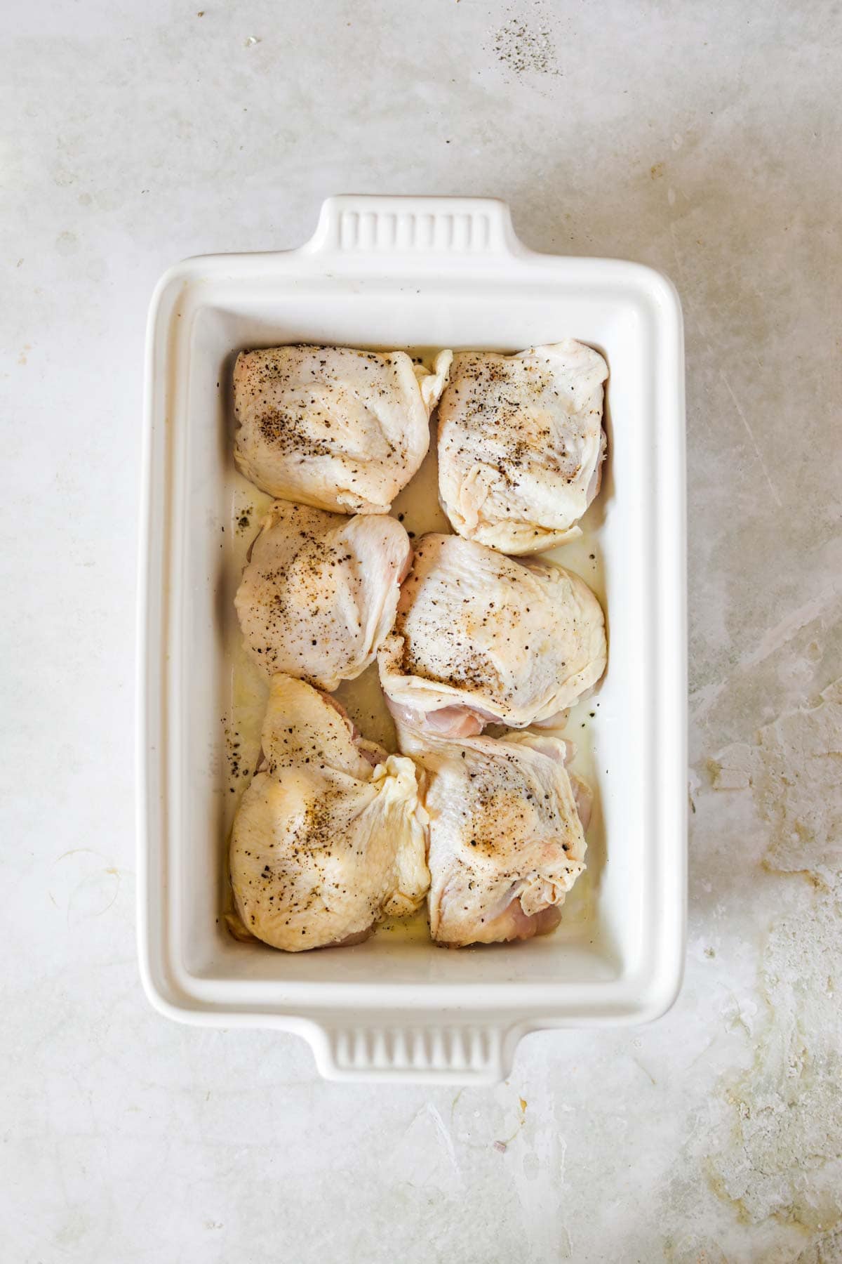 Seasoned chicken thighs in a white Le Creuset baking dish.