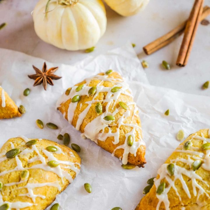 Pumpkin scones (better than Starbucks!) with maple vanilla paste glaze on parchment paper with decorative white pumpkins and cinnamon sticks.