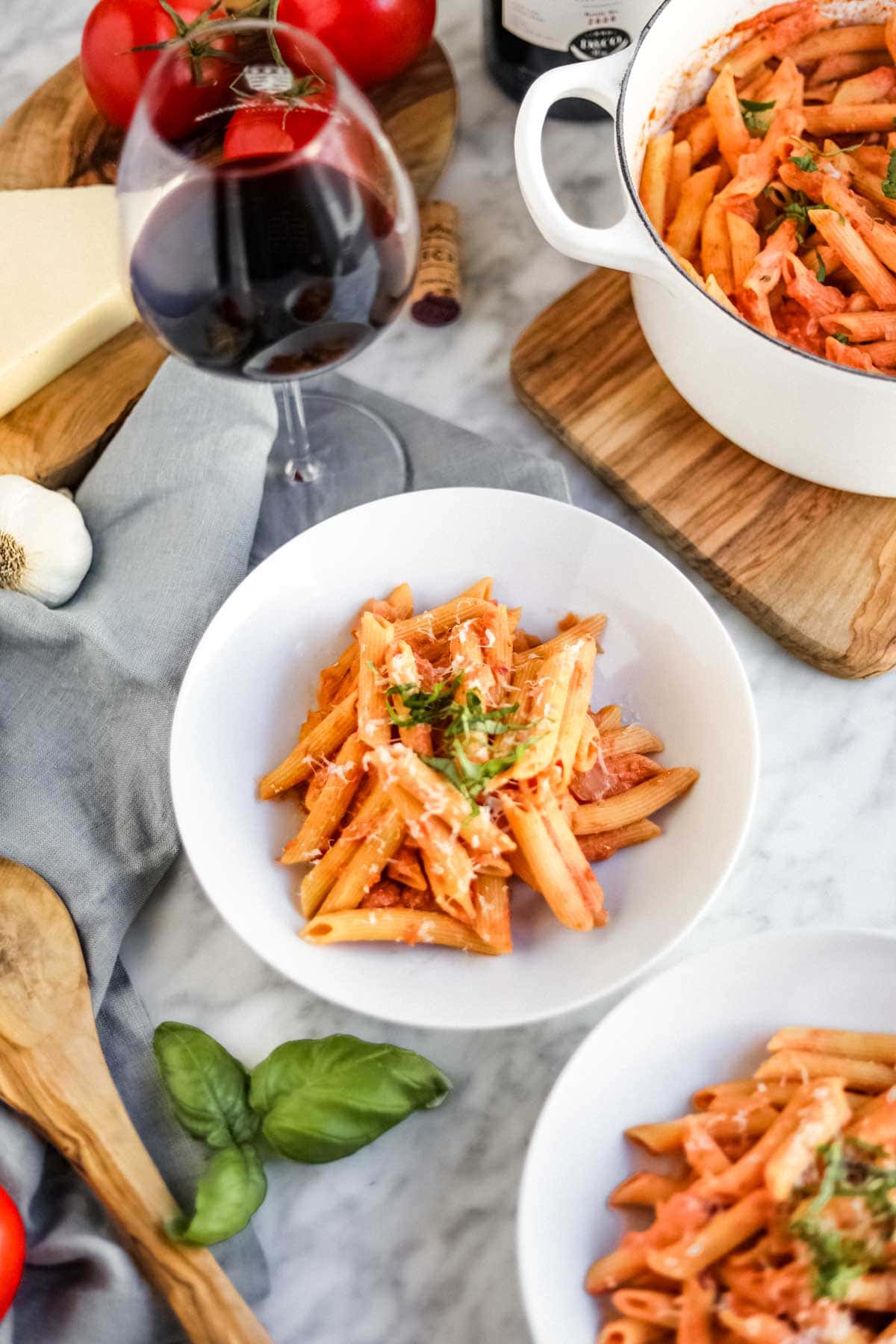 Penne pasta with vodka sauce in white bowls and dutch oven with wine, garlic, tomatoes and cheese in background.