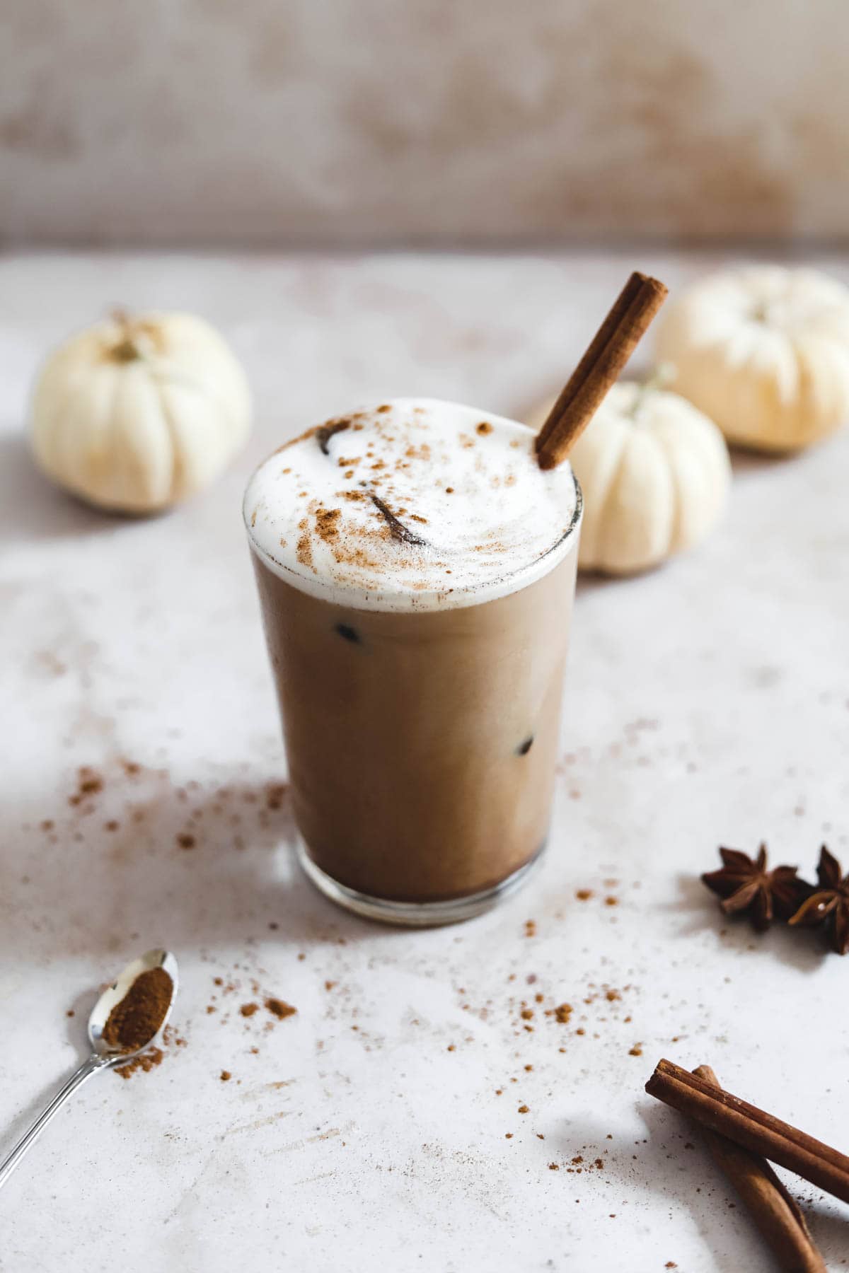 Iced pumpkin spice latte in a tall glass with coffee ice cubes, foam, spices, and a cinnamon stick for garnish and small white pumpkins, cinnamon sticks, and pumpkin spice as decor.