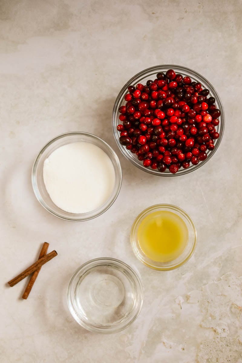 Ingredients for cranberry sauce in glass bowls, including fresh cranberries, orange juice, water, sugar, and cinnamon sticks.