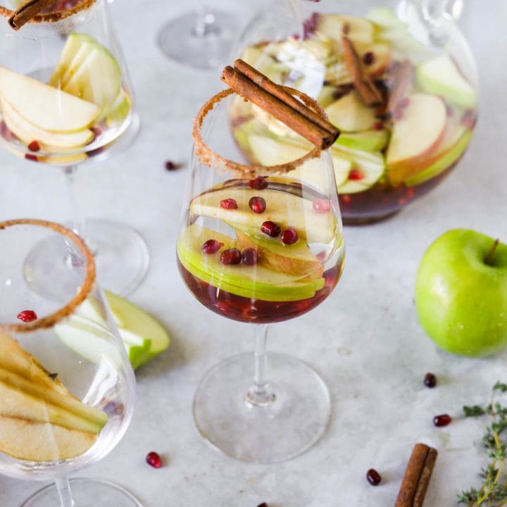 Fall sangria with pumpkin spice, apple, cinnamon sticks, and pomegranate seeds in a glass and a large glass pitcher.