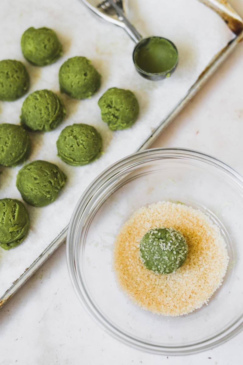 Green matcha cookie dough balls on a parchment paper-lined baking sheet and cookie dough rolled in coarse sugar in a bowl.