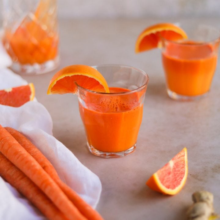 Carrot Orange Ginger juice in French glassware surrounded by fresh produce and sliced oranges.