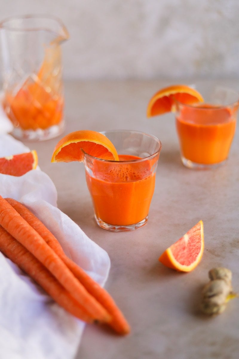 Carrot Orange Ginger juice in French glassware surrounded by fresh produce and sliced oranges.