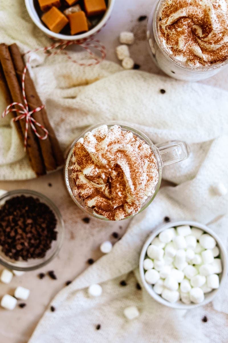 Flatlay image of oat milk hot chocolate in a mug with whipped cream and various toppings, including marshmallows, chocolate chips, and caramels.