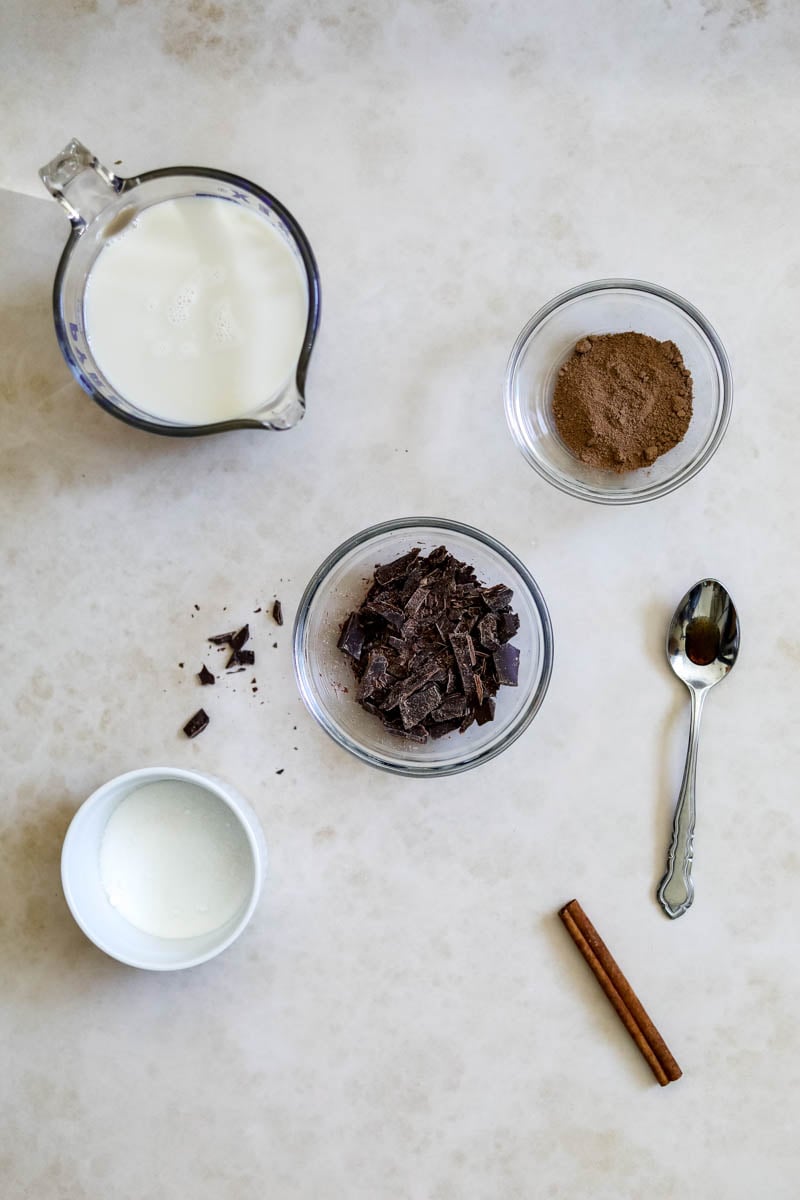 Ingredients to make oat milk hot cocoa, including oat milk, sugar, unsweetened cocoa, chocolate, cinnamon stick, and vanilla bean paste or extract.