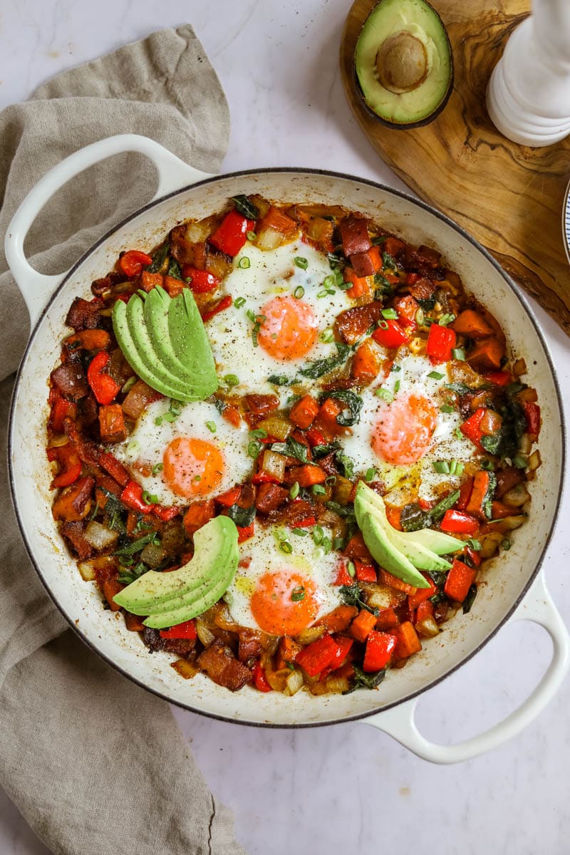 Breakfast hash with sweet potatoes, eggs, and avocado in a white Le Creuset braiser.