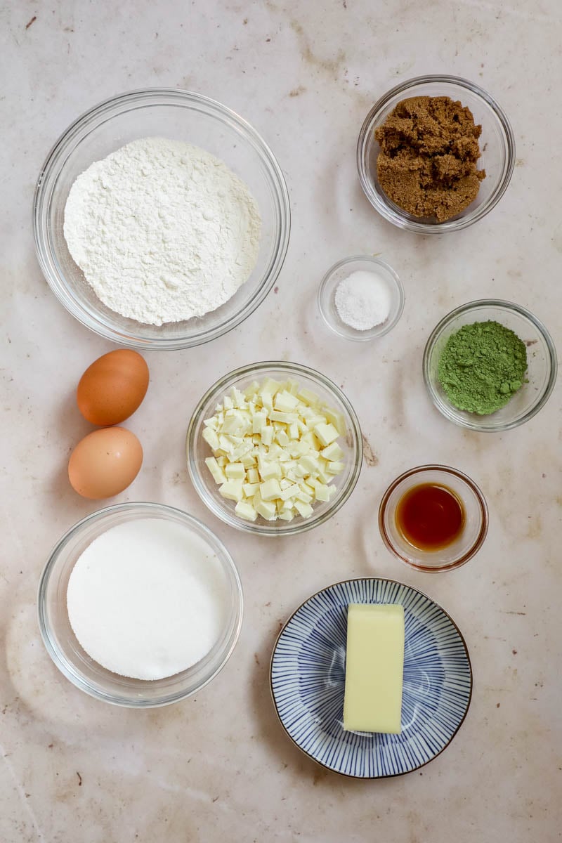 Ingredients in bowls to make matcha brownies (flour, eggs, sugar, white chocolate, butter, vanilla extract, brown sugar, matcha powder, and salt).