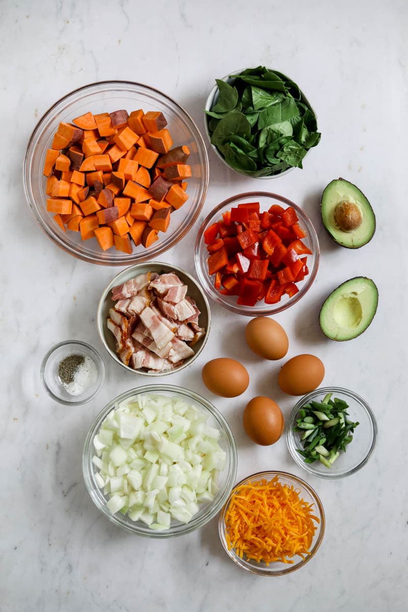 Ingredients for breakfast sweet potato hash (bell peppers, onion, cheddar cheese, eggs, avocado, green onion, salt, pepper, sweet potatoes, spinach).