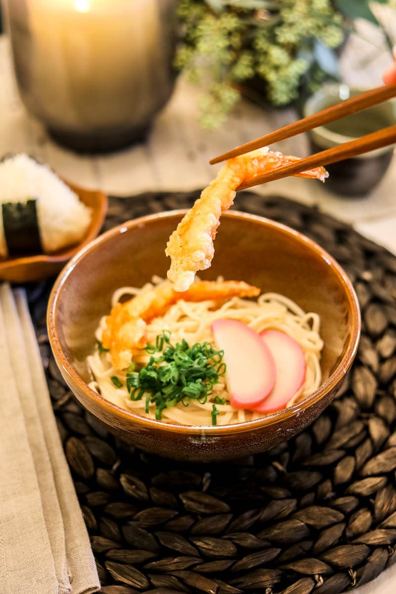 Shrimp tempura udon with chopsticks holding shrimp above noodles.