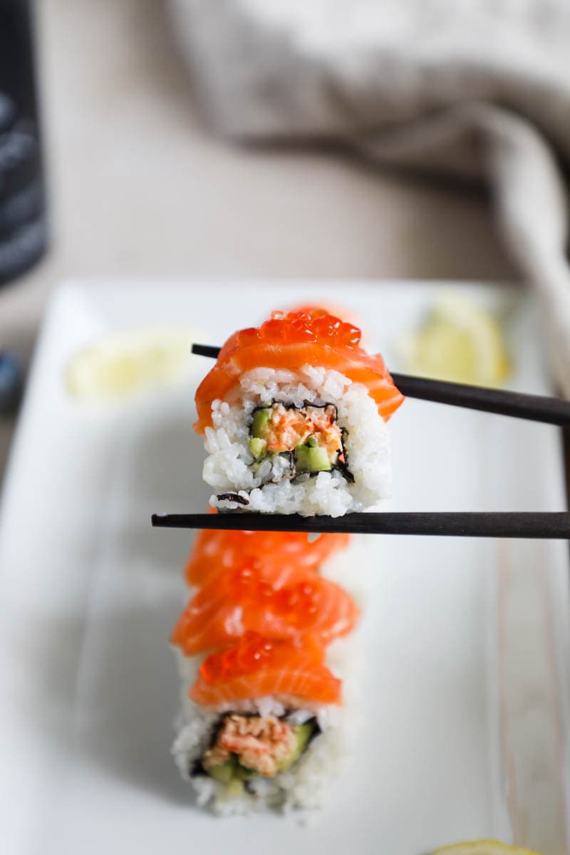 Alaska Roll held with chopsticks and topped with salmon and Ikura on a white plate.