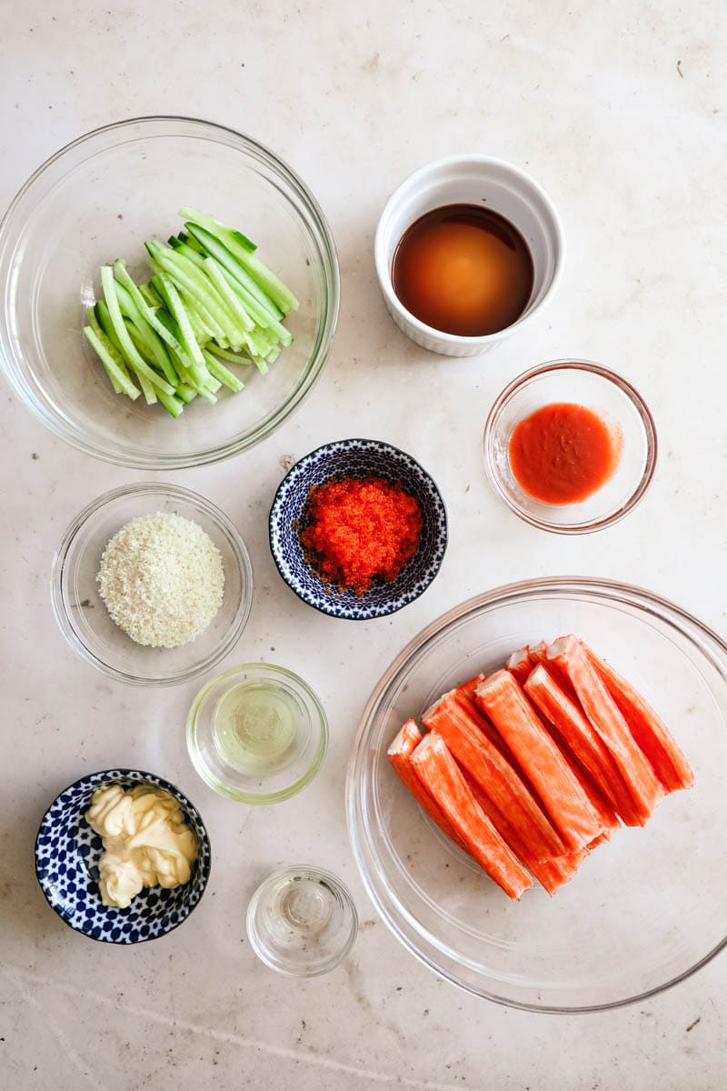 Ingredients to make kani salad, including sliced cucumber, imitation crab, tobiko, kewpie mayo, mirin, sriracha, soy sauce, and panko.