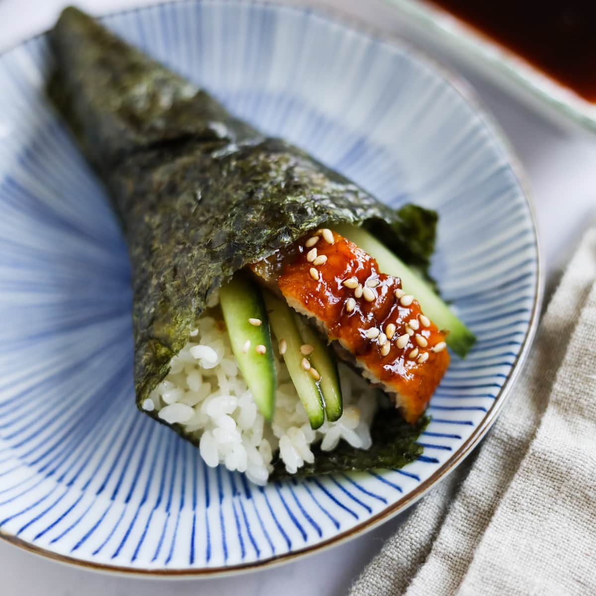 Unagi sushi (eel hand roll) with unagi, sesame seeds, and cucumber on a blue plate.