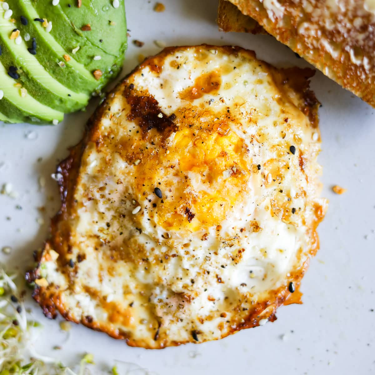 Over hard eggs with butter, salt, and pepper on a plate with avocado, toast, and sprouts.