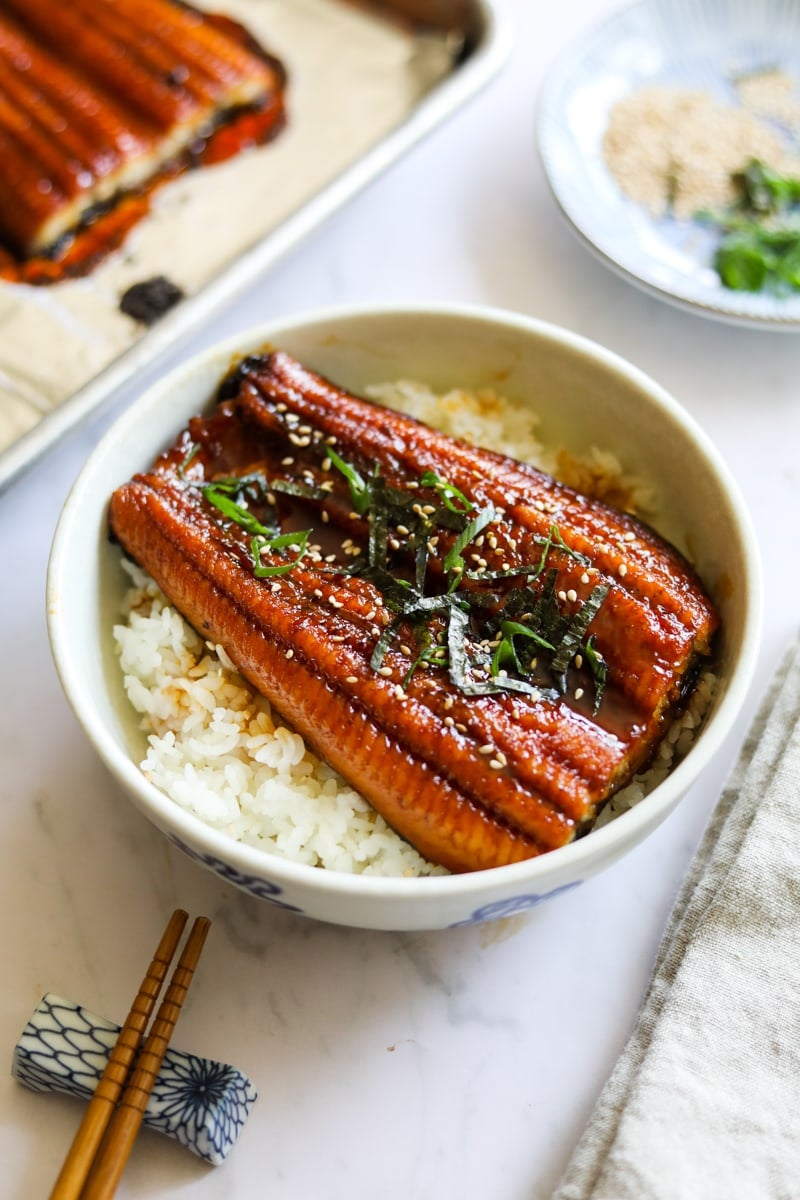 Unagi don (una don) in a white bowl with chopsticks, topped with unagi eel sauce, sesame seeds, nori, scallions on a bed of Japanese rice.