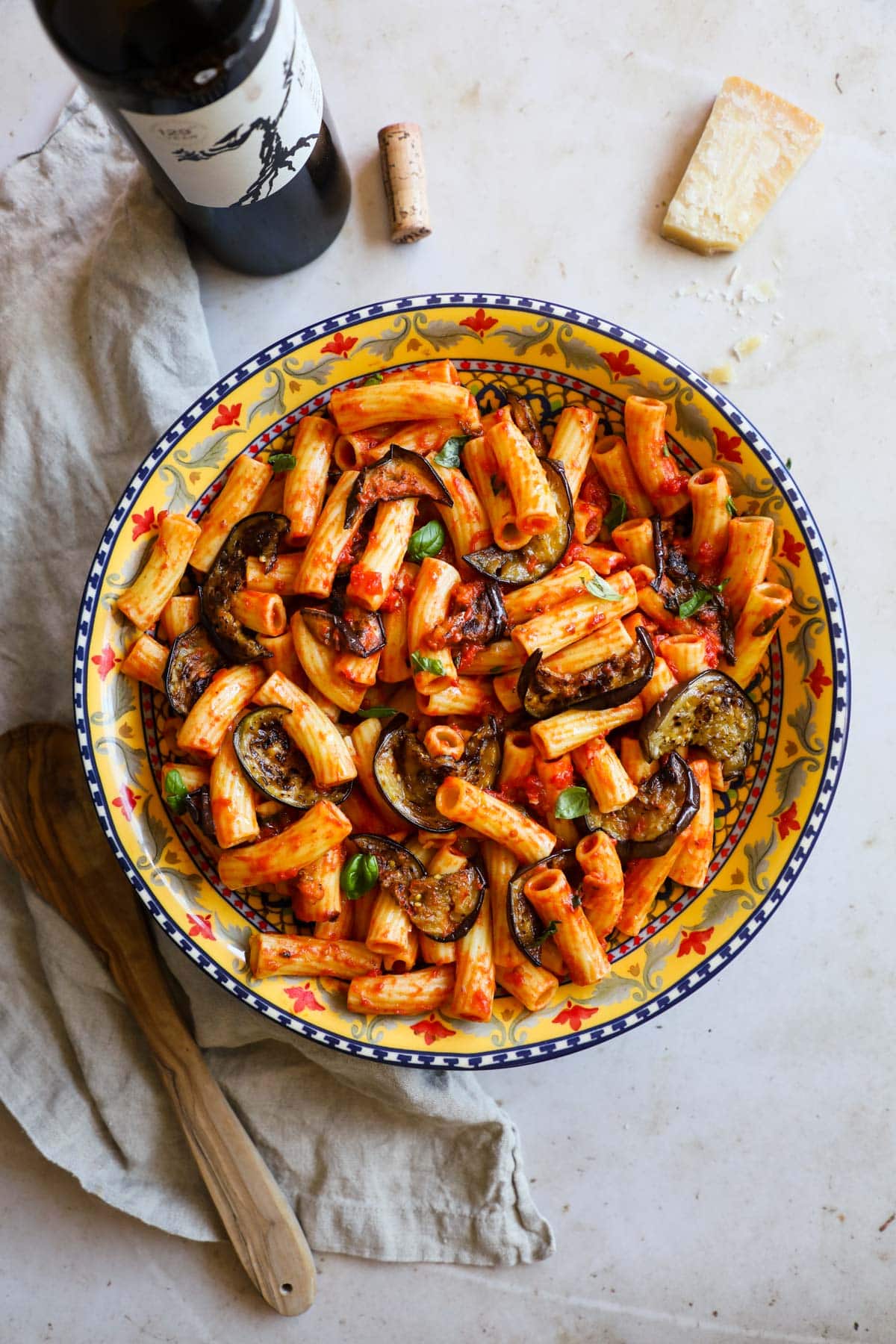 Eggplant pasta (pasta alla Norma) in a Sicilian ceramic bowl, Parmigiano Reggiano, wine, a linen napkin, and olive wood serving spoon.