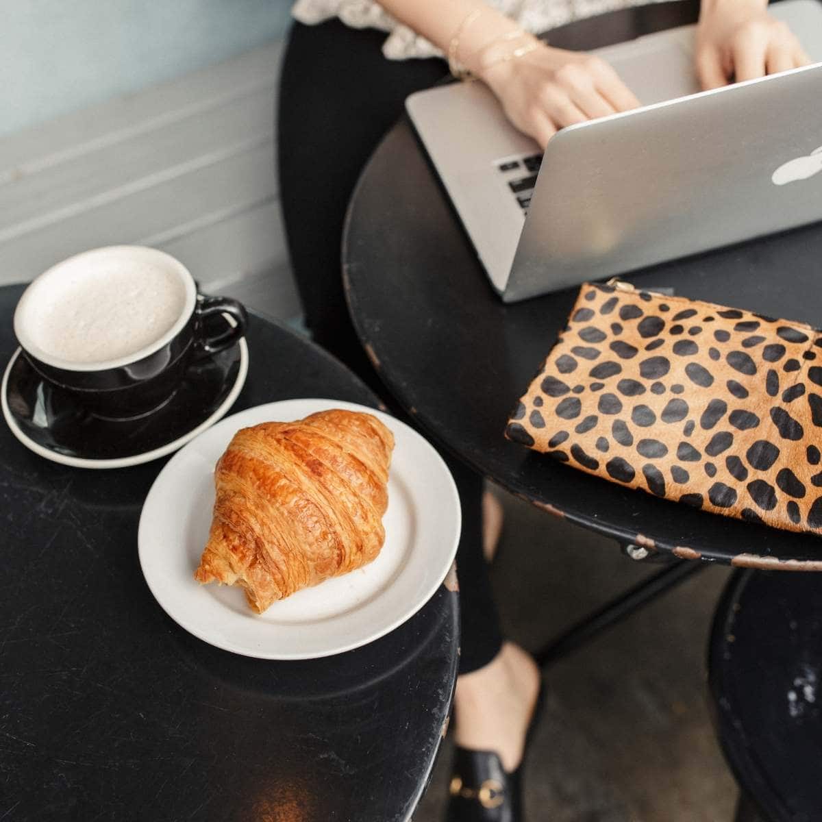 Kat typing on MacBook Pro at San Francisco cafe with croissant, latte, lace top, black jeans, and Gucci loafers.