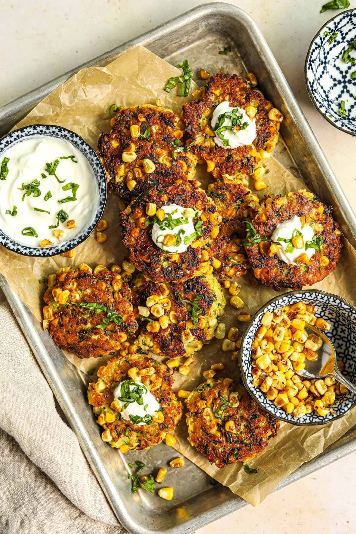 Overhead shot of healthy zucchini and corn fritters on a baking sheet with a bowl of greek yogurt and roasted corn for serving.
