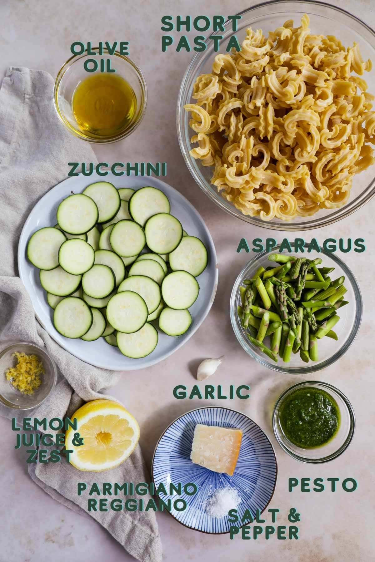 Ingredients to make veggie pesto pasta, including pesto, short pasta, olive oil, zucchini, garlic, asparagus, lemon zest and juice, and optional parmigiano-reggiano.