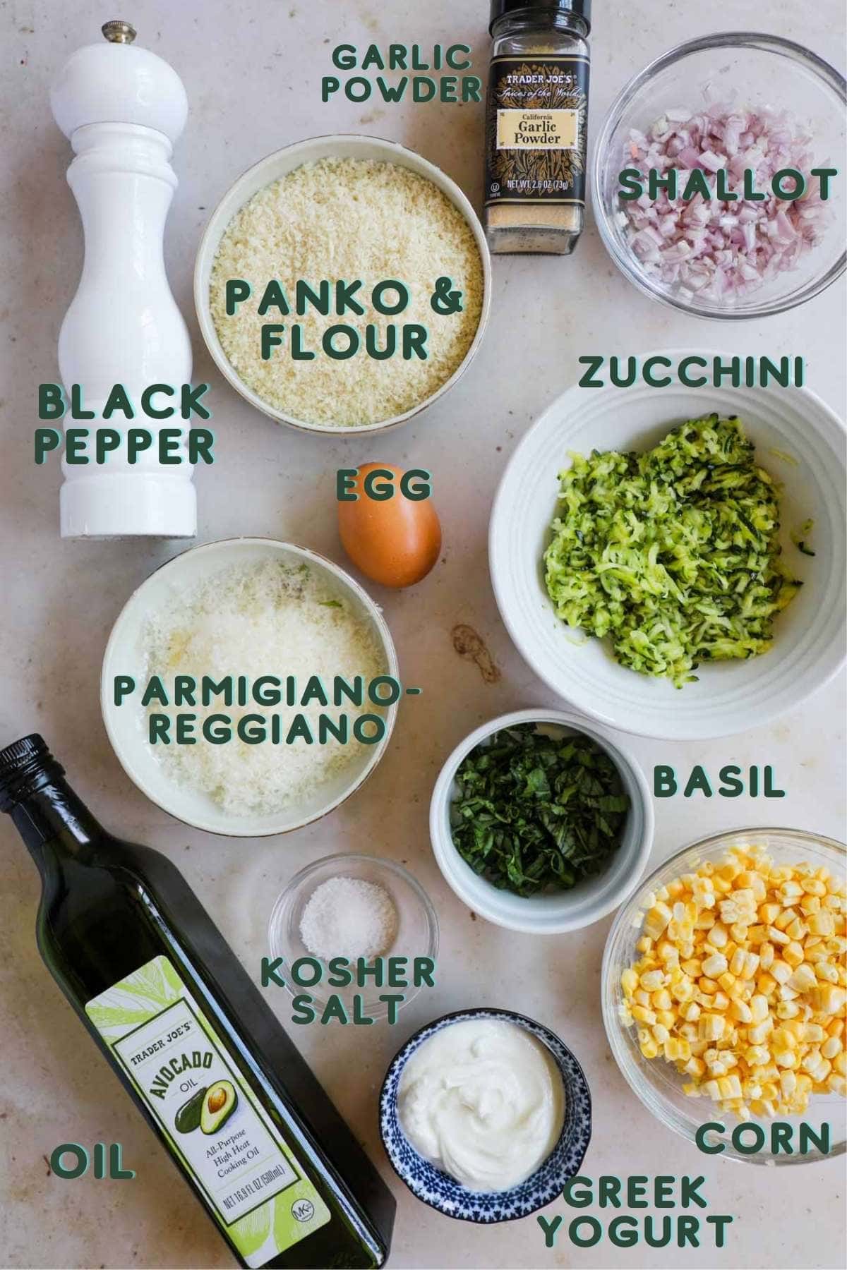 Ingredients to make corn and zucchini fritters, including grated zucchini, corn, parmigiano-reggiano, panko, flour, egg, garlic powder, shallot, basil, salt, avocado oil, greek yogurt, and black pepper.