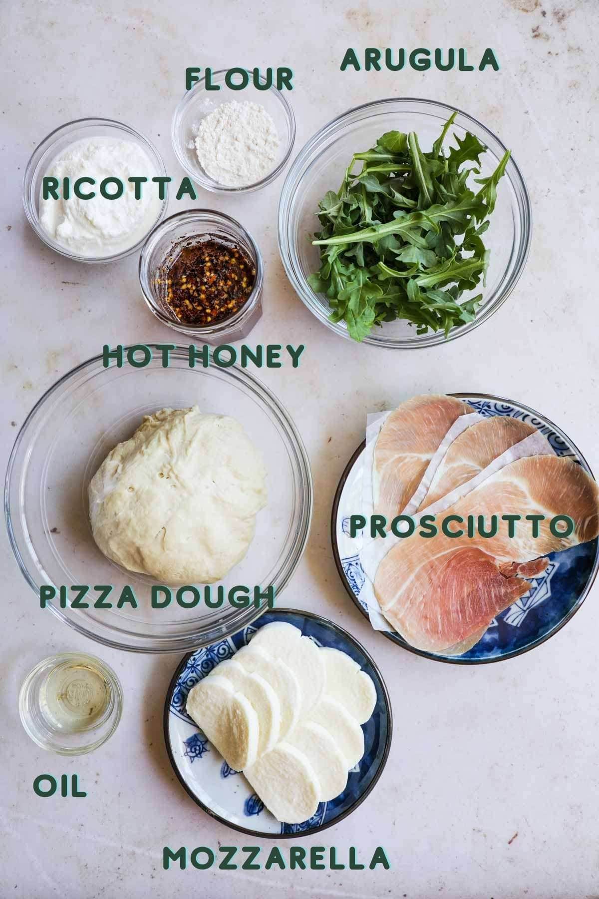 Ingredients to make pizza, including prosciutto, ricotta, arugula, pizza dough, mozzarella, flour, oil, and hot honey.