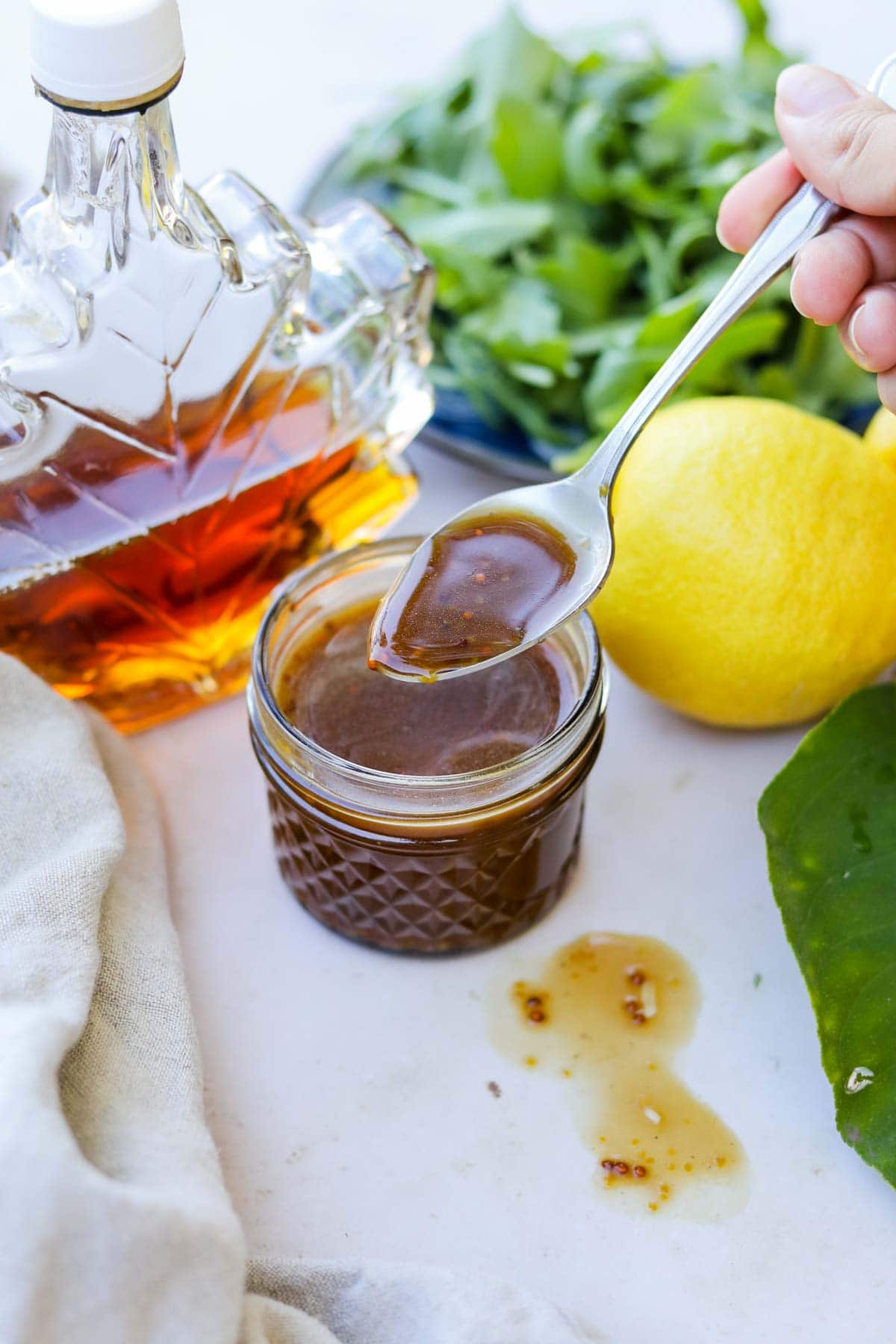 Spoon stirring maple balsamic vinaigrette dressing in a glass jar with maple syrup and lemon.