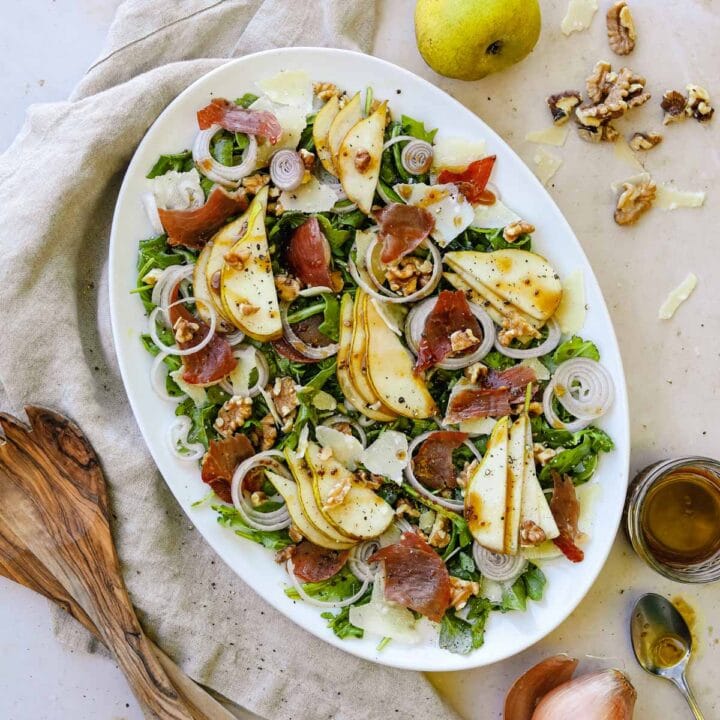 Pear and prosciutto rocket salad on a serving platter with sliced pears, crispy prosciutto, shallots, walnuts, arugula, parmigiano-reggiano, and a maple balsamic vinaigrette.