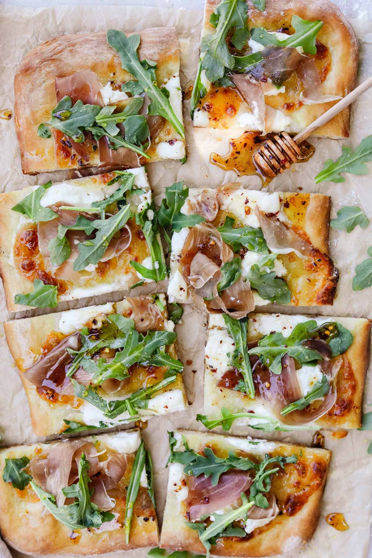 Overhead photo of pizza with prosciutto, arugula, ricotta, and hot honey on parchment paper with a honey dripper.