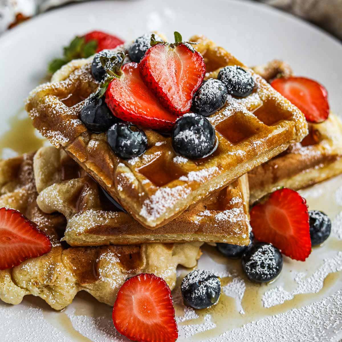 Gluten-free mochi waffles stacked on a plate with berries, maple syrup, and powdered sugar.
