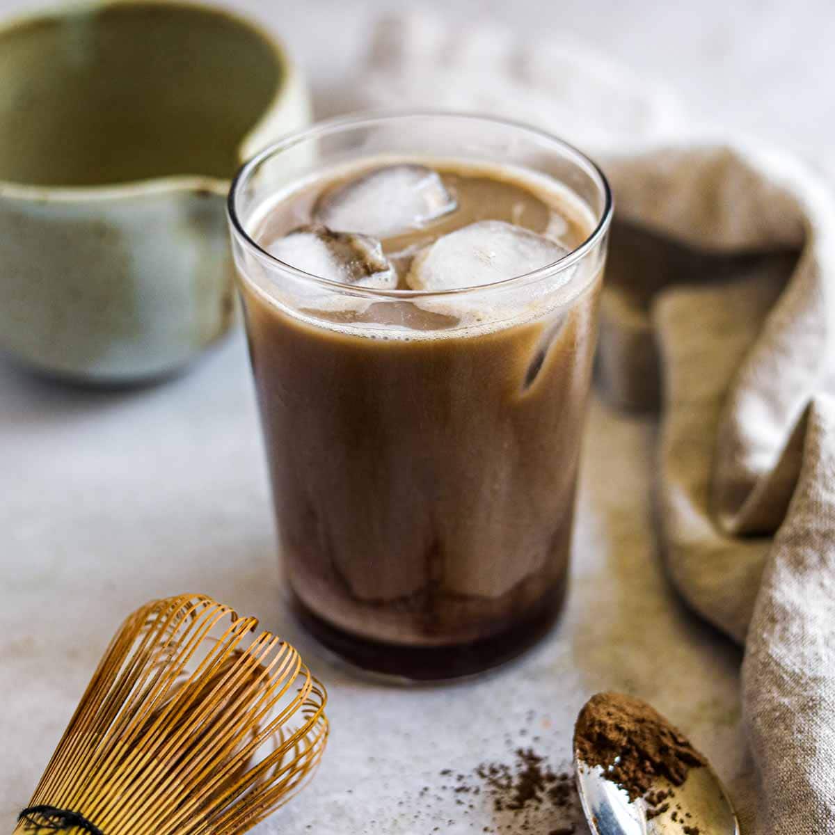 Iced Hojicha latte in a glass with ice, styled with a bamboo whisk and ground Hojicha.
