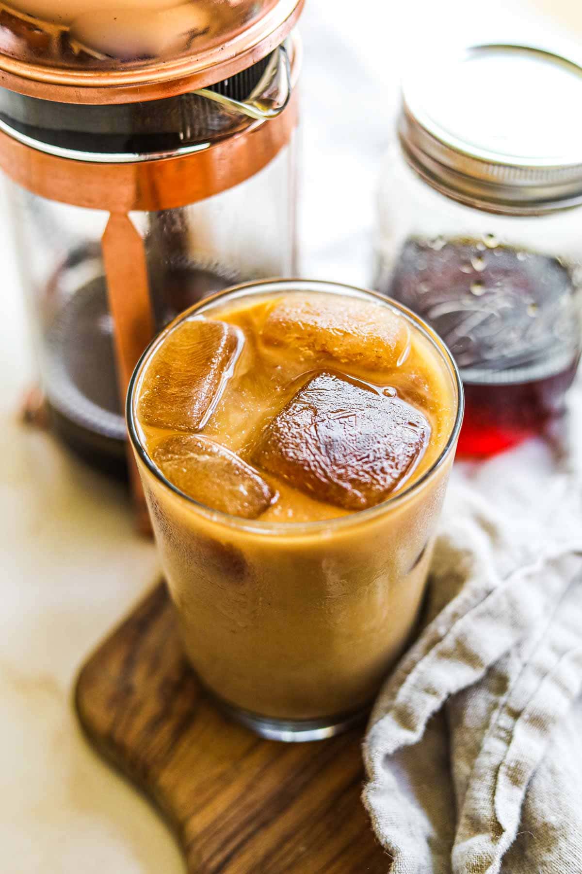 Caramel iced coffee in a glass with coffee ice cubes, a French press, and a jar of homemade caramel syrup.