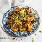Teriyaki fried tofu and Japanese eggplant in a homemade gluten-free honey teriyaki sauce topped with sliced green onions and sesame seeds on a blue plate.