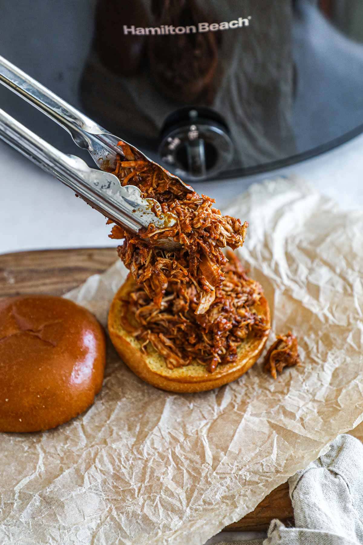 Tongs serving pulled slow cooker BBQ chicken thighs on a brioche bun.