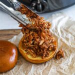 Tongs serving pulled slow cooker BBQ chicken thighs on a brioche bun.