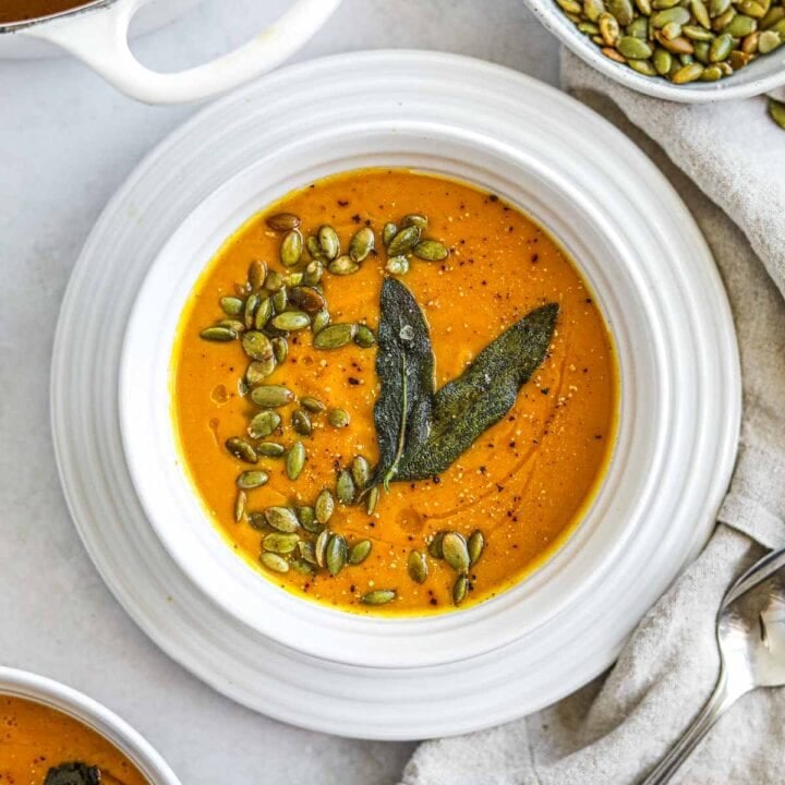 Vegan spiced butternut squash soup in a white Le Creuset bowl topped with roasted pepitas and fried sage leaves.