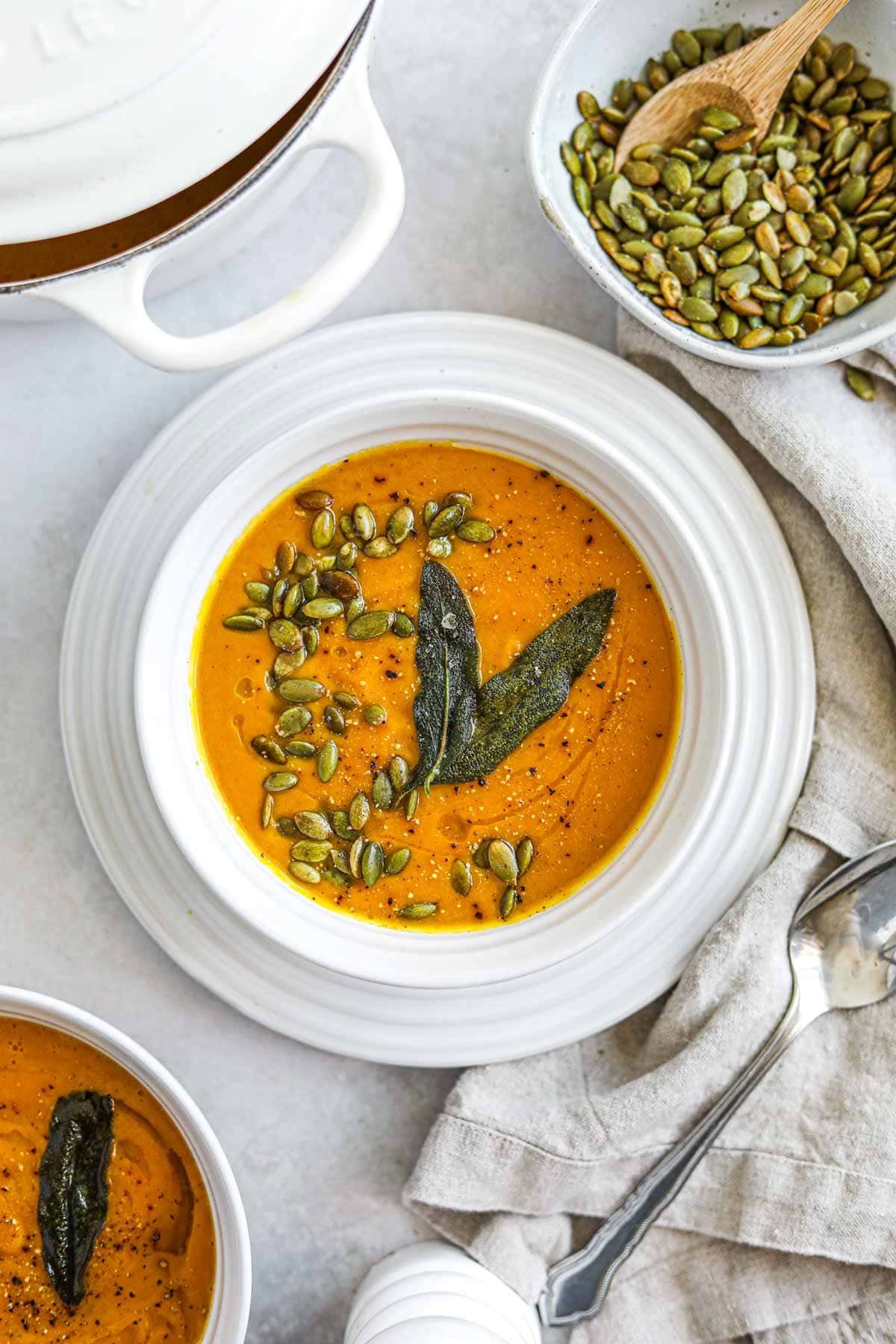Vegan spiced butternut squash soup in a white Le Creuset bowl topped with roasted pepitas and fried sage leaves.