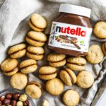 Baci di dama chocolate hazelnut cookie sandwiches in a basket with a jar of Nutella and a bowl of toasted hazelnuts.