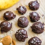 Frozen chocolate-covered banana bites with peanut butter and flaky sea salt on a parchment paper-lined baking sheet.