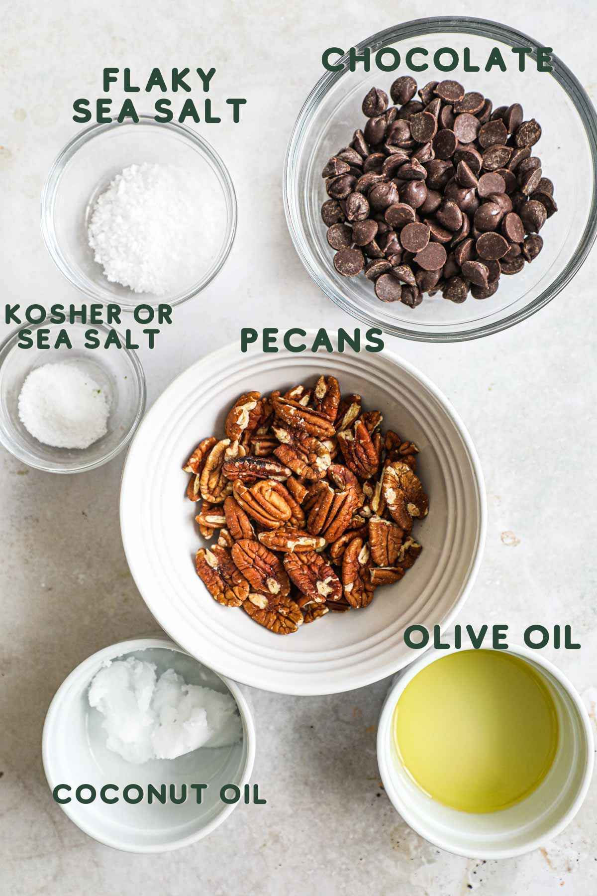 Ingredients to make homemade chocolate-covered toasted pecans, including chocolate, pecan, coconut oil, sea salt, flaky sea salt, and olive oil.
