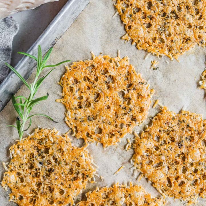 Montasio parmesan frico cheese crisps on a baking sheet with rosemary and black pepper.