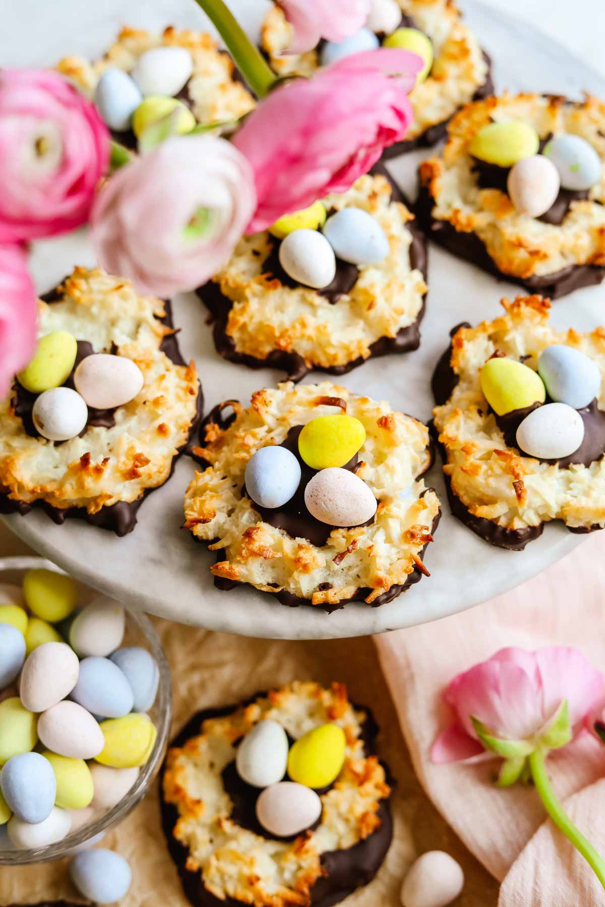 Chocolate-covered coconut macaroon nests with Cadbury mini eggs arranged on a marble platter with light and dark pink ranunculus flowers.