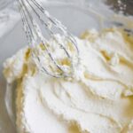 Fluffy, easy homemade whipped cream made with heavy whipping cream and sugar or honey in a chilled glass bowl with a mixer whisk.