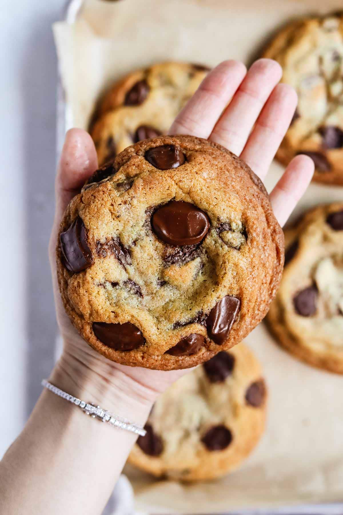 Hand holding a giant bakery-style chocolate chip cookie with crispy edges and a chewy center.
