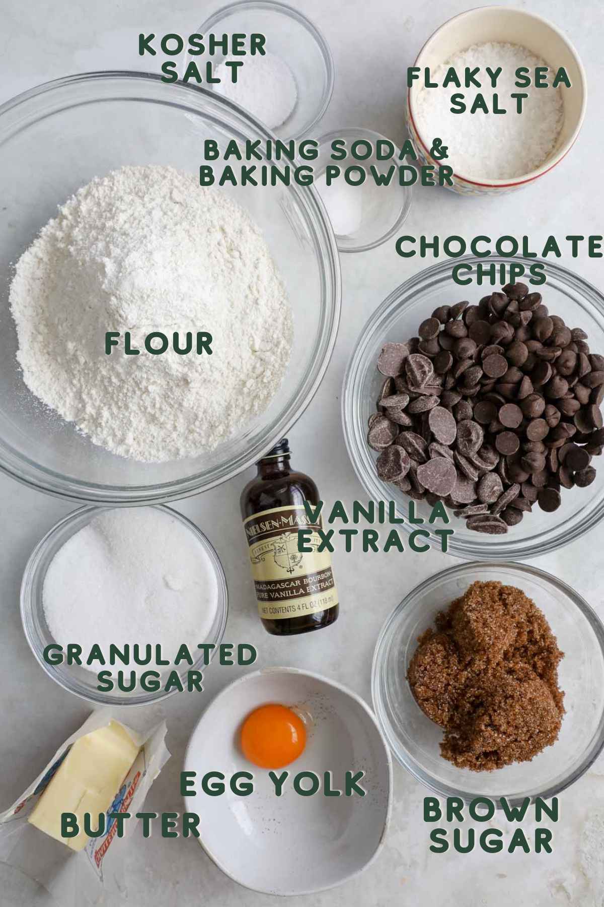Ingredients to make small batch chocolate chip cookies, including flour, vanilla, chocolate, egg yolk, butter, brown sugar, granulated sugar, baking soda and powder, and kosher salt.