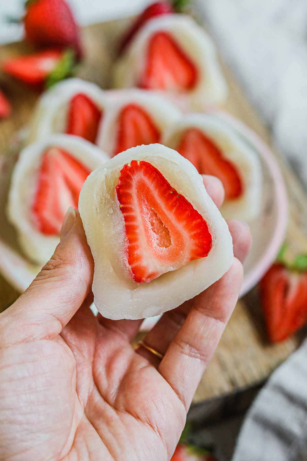 Hand holding a strawberry daifuku (mochi with bean paste) cut in half, revealing the strawberry and shiroan center.